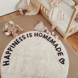A round, woven carpet with the text "Happiness is homemade" printed on it. The carpet appears to be a neutral beige color and is placed in a cozy, home-like setting with toys and other decor.