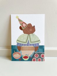 A whimsical illustration of a bear baking, surrounded by baking tools and ingredients. The bear appears to be wearing a chef's hat and apron, suggesting a birthday or celebration theme.
