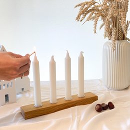 Tall, white candles on a wooden candle holder with a vase of dried flowers in the background. The candles appear to be made of wax and the candle holder is made of wood.