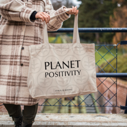  A neutral-colored or black tote bag with the text "Planet Positivity" printed on it. The bag appears to be made of a durable, canvas-like material and is being held by a person wearing a plaid jacket.