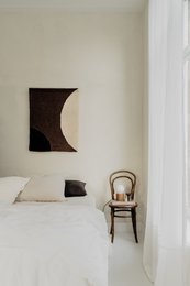Minimalist wall art with a crescent moon shape, made of dark material, mounted on a white wall. Wooden chair and couch with fur blanket in the room.