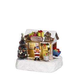 A miniature Christmas village scene with a small factory building, a decorated Christmas tree, and other festive elements. The scene has a cozy, whimsical feel with warm lighting and holiday decorations.