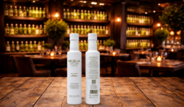  Two bottles of NIOELIA Extra Virgin Olive Oil on a wooden table in front of a bar with shelves of liquor bottles.
