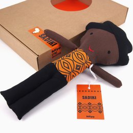 A plush African-inspired doll with a black body, orange and black patterned fabric, and a tag that says "Sadiki". The doll appears to be part of a baby doll collection.