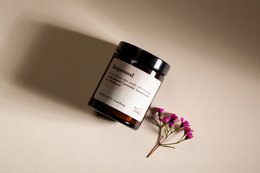 A round, tall, 155g scented candle in a glass jar with a black lid. The candle has a warm, amber-colored flame and is accompanied by a few small purple flowers.