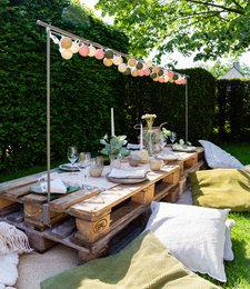 Outdoor string lights with colorful lanterns hanging over a rustic wooden table set for an alfresco dining experience in a lush garden setting.