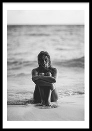 A black and white photograph of a young girl sitting on a beach, with the ocean in the background.