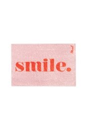 Plush pink bath mat with the word "smile" printed in large red letters. The mat appears to be placed on a wooden floor, with a person's feet visible in the foreground.
