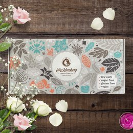 This deluxe gift box from Elly Momberg contains an assortment of sugar-free, vegan, and gluten-free chocolate products. The box features a floral design with various shapes and patterns, including leaves, flowers, and abstract elements. The products inside the box appear to be a variety of chocolate bars and other confections.
