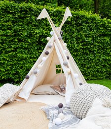 Outdoor light garland in a warm brown color, featuring a cozy teepee-style tent setup in a lush garden setting with string lights and decorative pillows.