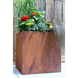 A large, square planter made of rustic, weathered wood. The planter is filled with a variety of lush, green plants including tall grasses and vibrant red flowers. The planter appears to be placed against a wooden fence, creating a charming, natural display.