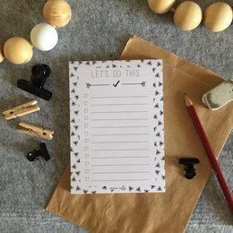 A white notepad with a patterned border, surrounded by various office supplies such as pencils, erasers, and a ruler. The notepad is titled "Let's do this" and appears to be part of the Notebooks category.
