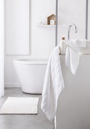  A pair of white, plush towels hanging on a towel rack in a minimalist, white bathroom with a freestanding bathtub.