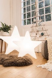 A large, illuminated white star-shaped decoration sits on the floor in a cozy, rustic-style room with a large window. The star appears to be made of a soft, plush material and is surrounded by other decorative elements like a small potted plant.