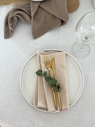 Muslin napkins in a variety of neutral colors including white, beige, taupe, light gray, dark gray, light blue, rose wood, blush, champagne, khaki, indigo, blush pink, sage, and mint. The napkins are shown on a table setting with a fork and knife.