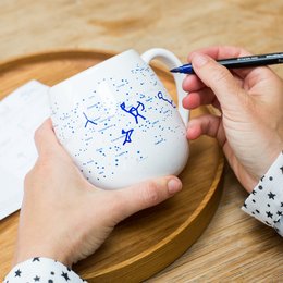 A white ceramic mug with a constellation pattern that can be colored in using the included pen. The mug is designed for creative expression and personalization.