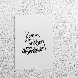 Postcard with handwritten text "Komm, lass uns ein Abenteuer!" in black on a white background. The postcard appears to be a Christmas or holiday-themed item.
