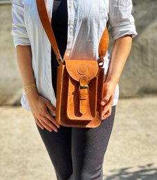  Handmade genuine leather crossbody bag in a rectangular shape with a flap closure and a buckle strap. The bag has a warm brown color and a vintage style.