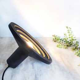 A round, tall table lamp with a textured, stone-like surface in shades of beige and brown. The lamp has a warm, glowing light that creates a cozy, natural ambiance.