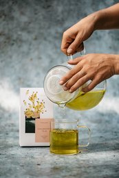Loose tea in a glass jar with a wooden lid, accompanied by a glass container of golden liquid, likely oil or honey. The tea appears to be a light green or yellow color, suggesting a delicate, floral variety such as cowslip tea.
