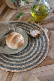 Circular jute placemat in neutral tones of cream, pink, olive, and grey. Placemat features a woven, textured design and is paired with a round bread roll and olive oil.