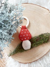  A small, round mushroom-shaped rattle with a red polka dot cap and a white stem, nestled on a wooden slice surrounded by greenery.