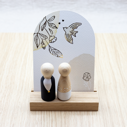 "Decorative wooden figurines depicting a bride and groom, displayed on a wooden stand with a floral illustration and the word 'Liefde' (Dutch for 'love'). The figurines have a simple, minimalist design."