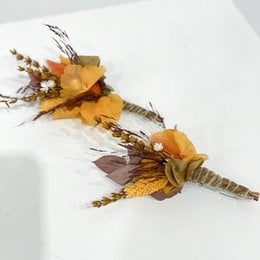 A brooch with dried flowers and leaves in warm autumn colors, including orange, yellow, and purple. The brooch has a rustic, natural appearance and would be suitable for formal or casual wear.