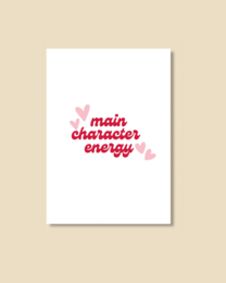  A pink and red heart-shaped postcard with the text "main character energy" printed on it. The postcard is placed on a wooden surface.