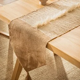 A long, rectangular table runner made of beige, woven material with a textured, natural appearance. The runner is designed to be placed on a table, adding a rustic, earthy touch to the decor.