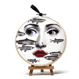 The product is a wall circle featuring an embroidered design of the face of Lina Cavalieri, a famous Italian opera singer. The design has a surreal, artistic style with bold features and expressions. The product is part of a collection and available in several variants, including Lina smoke, Lina 2 profile, Lina pirate, Lina Clown, and Lina terrified. The wall circle measures 15 cm in diameter and would make an eye-catching and unique decorative piece for the home.