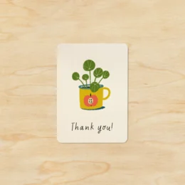 A small, rectangular thank you card featuring a green plant in a yellow watering can. The text on the card reads "Thank you!"