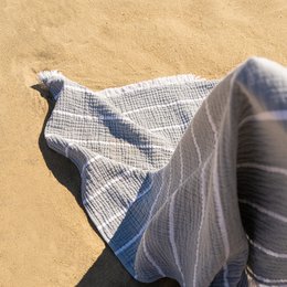 A large, gray and white striped peshtemal or hammam towel, folded and displayed on a wooden chair or bench against a sandy background.