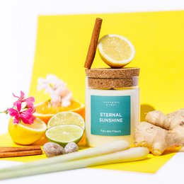  A tall, round, yellow scented candle with a cork lid and a lemon slice on top, surrounded by fresh ginger, lemon slices, and pink flowers on a yellow background.