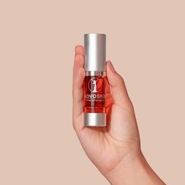 Antiaging eye serum in a red bottle with a silver cap, held in a hand against a beige background.