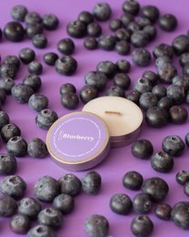 Small round blueberry scented soy candles in a purple container, surrounded by fresh blueberries on a vibrant purple background.