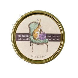 A round, gold-rimmed candle with a whimsical illustration of a rabbit and various stationery items. The candle is labeled "Tokyomilk" and appears to be a scented candle product.