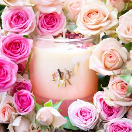 A round, tall candle with a rose and vanilla scent, surrounded by pink and peach roses.