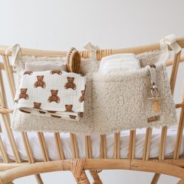A baby crib organizer in cream and taupe colors, featuring a teddy bear design and additional storage compartments for diapers and other baby essentials. The organizer is made of soft, plush materials and attaches securely to the crib for convenient access.