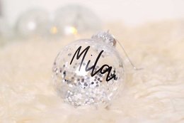 Clear glass Christmas ornament with the name "Mila" written on it, decorated with glitter and sparkles.