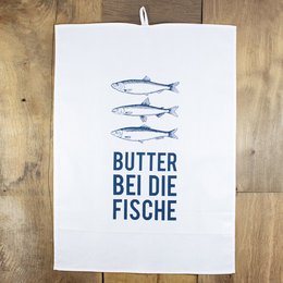 White tea towel with blue text "Butter bei die Fische" and three illustrated fish. The tea towel appears to be made of cotton and has a hanging loop.