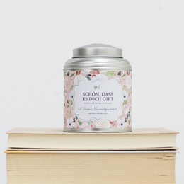  A floral-patterned tin canister containing a tea blend with the text "It's nice that you exist" printed on the label. The canister is placed on a wooden surface, with a woman's hand holding a similar canister in the background.