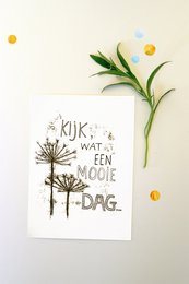 A white card with handwritten text that says "Kijk wat een mooie dag" (Look what a beautiful day) surrounded by a floral arrangement of white flowers and green leaves.