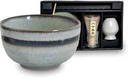 A ceramic matcha bowl set in a gift box, including a matcha whisk and scoop. The bowl has a mottled gray and white glaze, giving it a rustic, earthy appearance. The set is designed for preparing and serving traditional Japanese matcha tea.