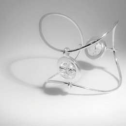 A large, chunky cuff bracelet made of silver-toned metal with two large, round gemstones - one smoky quartz and one rock crystal. The bracelet has a bold, modern design with a sleek, minimalist aesthetic.