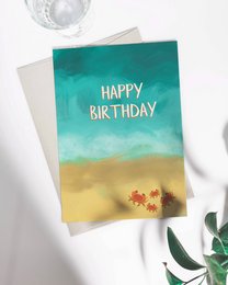 A colorful birthday card featuring a beach scene with a crab. The card has the text "HAPPY BIRTHDAY" prominently displayed.