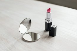 Compact round pocket mirror with a red lipstick. The mirror is made of metal and has a sleek, modern design. The lipstick is a vibrant red color.