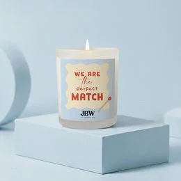 Cream white cylindrical scented candle with "We Are The Perfect Match" text and decorative matchstick design, JBW brand.