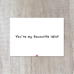A white greeting card with the text "You're my favorite idiot" printed on it, against a wooden background.