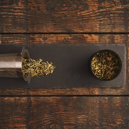 A black tea infuser with a metal mesh filter and a round tea strainer made of gold-colored metal, both placed on a dark wooden surface.